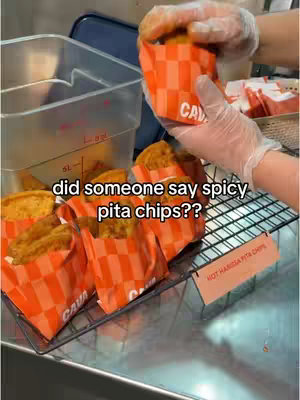 A person wearing gloves is holding a bag of pita chips next to a display of similar bags and a container. The bags are orange and white checkered, with 'CAVA' printed on them. A sign reads 'HOT HARISSA PITA CHIPS'.