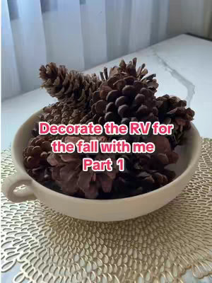 The image is an overhead shot of a large, cream-colored bowl overflowing with brown pinecones set on a gold, patterned surface. The background shows glimpses of white curtains and a white surface. In the center, a text overlay in pink letters reads, "Decorate the RV for the fall with me Part 1."