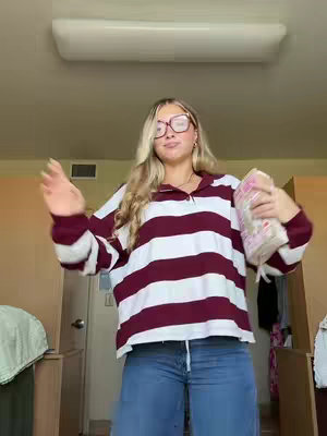 A woman with long blonde hair and glasses is gesturing with her right hand towards the audience. She is wearing a maroon striped shirt with blue jeans. In her left hand, she is holding a pink bag with what appears to be product packaging.