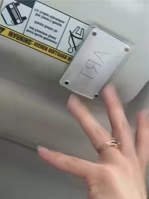 A hand with a ring is seen in the foreground, reaching towards a rectangular object attached to the ceiling. The object has stylized text on the side facing the camera. Above this and to the left are warning labels with text and a car icon.