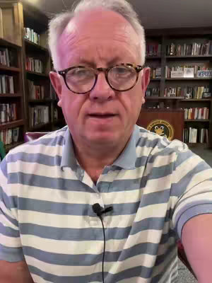 A man with glasses and a striped shirt is speaking to the camera. He is in front of a bookshelf and is lit by overhead lighting.