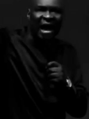 A grayscale image features a man with a focused expression, possibly in the middle of speaking passionately. His face is the central focus, showing him from the chest up, with his dark clothing adding a stark contrast against a darker background. The lighting emphasizes the emotional intensity of the moment, common in impactful speaking engagements.