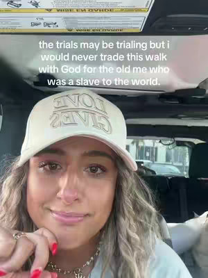 A young woman is seated inside a vehicle, likely a car or SUV, with a portion of the ceiling visible above. Her head is in the center of the frame against a neutral background, she's wearing a cap with the words "ONE GRACE" on it. The text overlay that reads "the trials may be trialing but I would never trade this walk with God for the old me who was a slave to the world."She's looking downwards, with an expression that may be introspective or slightly emotive.