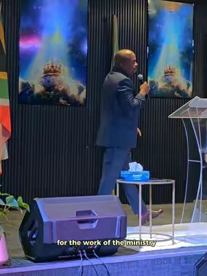 The image shows a man in a blue suit walking to the left while speaking into a microphone. Behind him are two large, vertical abstract paintings with a blue and gold-like color scheme. There's a small table with tissues on it and a black speaker in the lower left.