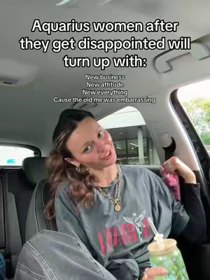 A woman is seated in a car, slightly angled toward the camera. She has light skin and is wearing a black beanie, a gray t-shirt with a graphic, gold jewelry, and a black top. Her hair is styled away from her forehead. The image has overlay text that presents a meme.