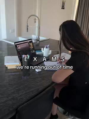 A medium shot shows a woman sitting at a kitchen island, studying from a laptop. Text overlays read "EXAMS" and "we're running out of time." The setting appears to be a modern kitchen with a stainless steel sink.