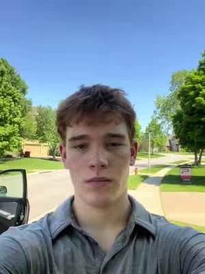 A selfie of a young man in a gray shirt stands outdoors on a sunny day, with a street and houses visible in the background.