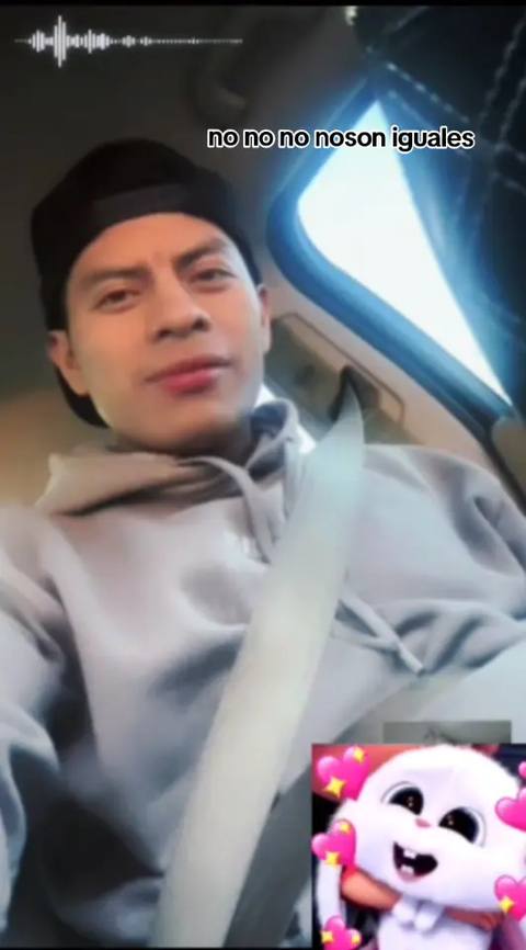 A selfie-style video taken from inside a car. The main subject is a young man wearing a black cap and a gray hoodie, looking at the camera. He has a neutral expression.  In the bottom corner, there's a small animated image of a white bunny with pink hearts. Behind him, the car's interior and the outside world are slightly visible creating depth.