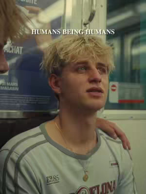 A young man with short, messy blonde hair is the central focus, his face conveying a mix of distress and contemplation. He's wearing a light gray t-shirt with a sporty design and a gold necklace. An arm is seen to his right, offering a gesture of support or comfort. The background appears to be inside a vehicle, possibly a subway train, with advertisements or signs barely visible. The color palette is muted, with cool tones that highlight the man's facial expression.