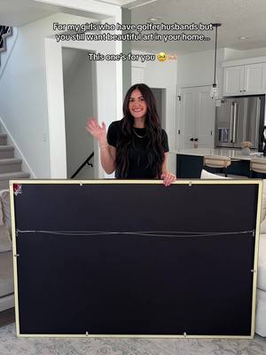 A woman holding a large framed artwork with a black background, standing in a well-lit room. She is smiling and waving at the camera. The background shows a modern kitchen and living area with stairs. Text overlays at the top.