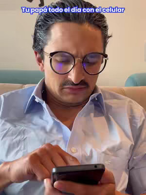 A middle-aged man with dark hair, round glasses, and a mustache is looking down at a smartphone. The text overlay reads "Tu papá todo el día con el celular."