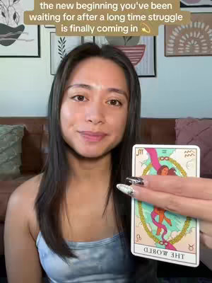 A woman with long dark hair and a light blue tank top is shown from the chest up, holding a tarot card that displays an image of a woman surrounded by a garland. Behind her, a brown couch and framed artwork are visible. Above her head, text is overlaid in a rectangular shape.