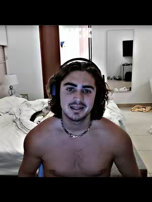 A shirtless man with dark hair and headphones sits in front of a bed, likely in a bedroom setting. The lighting appears dim or natural, creating a somewhat intimate atmosphere.