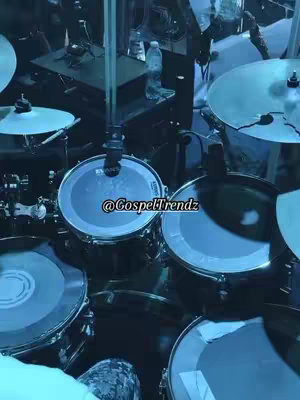 An overhead view of a drum set. The drums are black and silver, and the cymbals are silver. The overall tone is dark and moody. The angle is from behind the set looking forward. Text "@Gospeltrendz" is added to the drum heads.