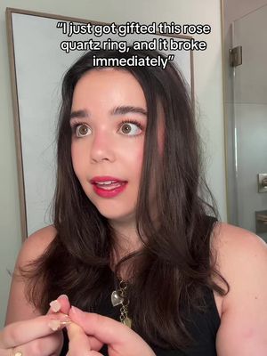 A selfie-style image of a woman with long, dark hair, looking directly at the camera with wide eyes and a slightly open mouth, as if surprised or shocked. The background is a light-colored wall with a portion of a mirror visible. Text overlayed at the top states her surprise at a ring breaking.