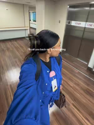 A woman with dark hair in a bun, wearing blue scrubs, a pink name tag, and a backpack, is walking down a hospital corridor. The background includes a light-colored hallway with a wooden floor and closed doors. Text overlayed says, 'I'll call you back my future calling.'