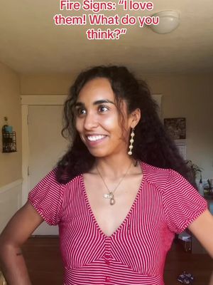 A woman with long, curly dark hair stands in a well-lit room, smiling. She's wearing a red and white striped top and gold jewelry. Text on top reads: 'Fire Signs: "I love them! What do you think?"'
