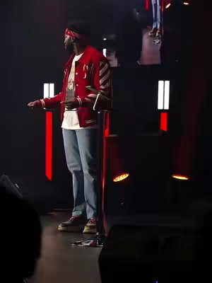 A person stands on a stage in front of a dark background with vertical illuminated rectangles. The person, wearing a red jacket, jeans, and a red bandana, gestures as if speaking.