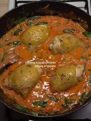 A top-down shot of a pan filled with creamy Tuscan chicken. The dish features large, golden-brown chicken pieces submerged in a thick, orange-red sauce dotted with bits of green. The pan sits on a stovetop, indicating a home-cooked meal. Text overlays indicate the name of the dish.