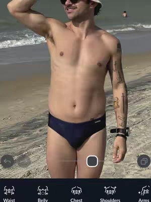 A man in sunglasses and hat standing on a beach. He is shirtless and wearing dark blue swim trunks. He is holding his arm above his head, and there are icons at the bottom of the picture.
