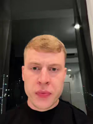 A close-up shot of a man's face, positioned in the center of the frame. He has short, light-colored hair. The background is dark, out of focus with a city view visible outside a window. The man is looking directly at the camera.
