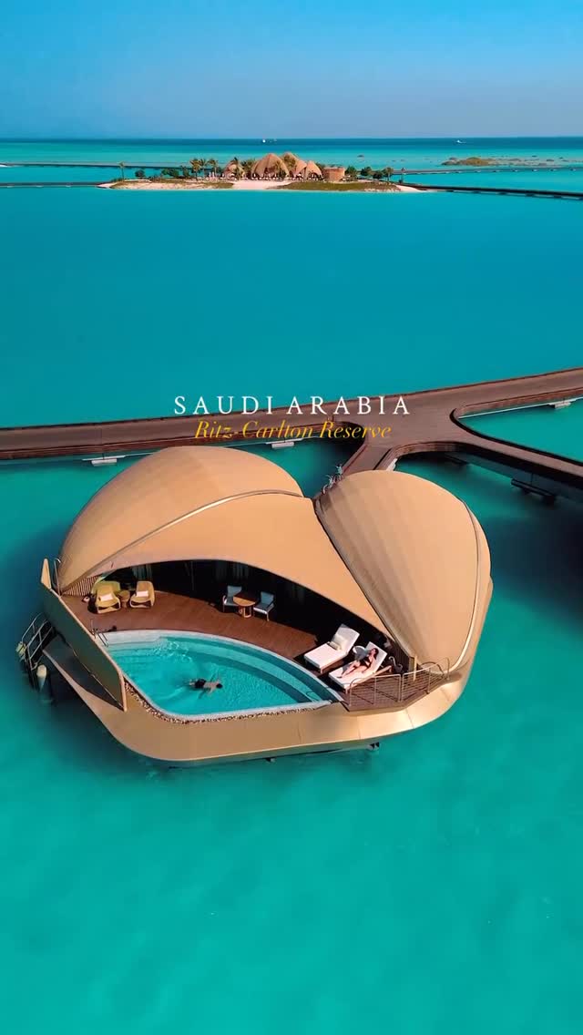 An aerial view of an overwater villa in a turquoise sea. Brown bridge leads to floating villas that have a pool and beach cabana, in the background there are more hotels and resorts.