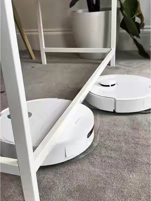 The image shows two white robot vacuums on a gray carpet. A white metal frame of a table is in the foreground, partially obscuring the view. In the background, a white plant pot and a snippet of a plant are visible, and the wall is painted gray.