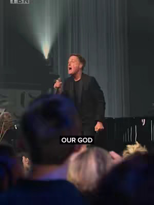The image captures a live performance. A man, center stage, sings into a microphone with conviction. He's dressed in a dark blazer. A stage backdrop and lighting elements provide context. A partial view of the audience is visible, adding to the live performance atmosphere. Text overlay reads 'OUR GOD'.