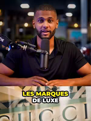 A man with a beard and short dark hair is centered in the frame, speaking into a microphone. Behind him, a blurred background includes dark, possibly indoor, setting. In the bottom portion of the frame, just underneath the man, is a decorative sign with the word "GUCCI" on it. The overall composition is a medium shot, well-lit, and the man appears to be the focus, centered with a neutral expression.