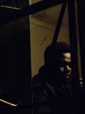 The image shows a dark, low-lit environment. The main visual element is a person, partially visible through a window frame. The person's face is in shadow, with only some features discernible. The lighting suggests a nighttime setting. The composition is simple, with the window frame and the person's silhouette dominating the space.