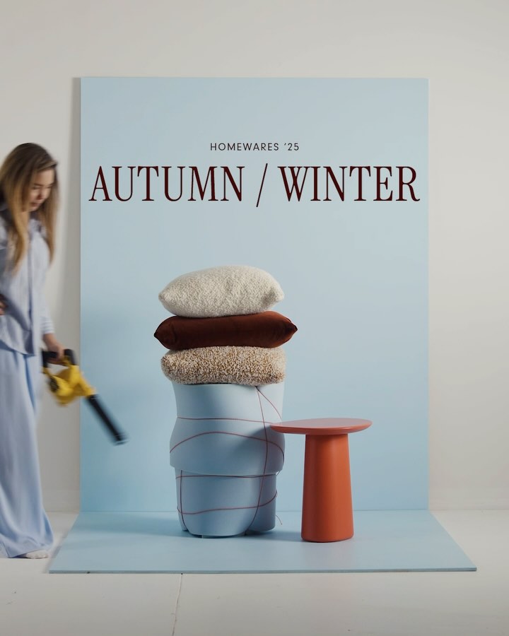 A studio shot featuring a light blue background with a stack of pillows of varying textures and colors. A woman in light blue clothing is entering the frame from the left, holding a yellow leaf blower. The text "HOMEWARES '25" is above the text "AUTUMN / WINTER". To the right, there's an orange side table. The overall composition is modern, clean, and incorporates autumnal colors.