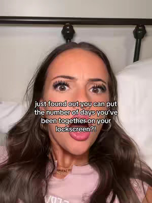 A selfie-style shot featuring a woman with dark hair in a pink top, lying in bed. Text overlay details a feature regarding the number of days a couple has been together on their lockscreen. The woman has expressive features, with a surprised or excited facial expression. The background includes a bed frame and white bedding.