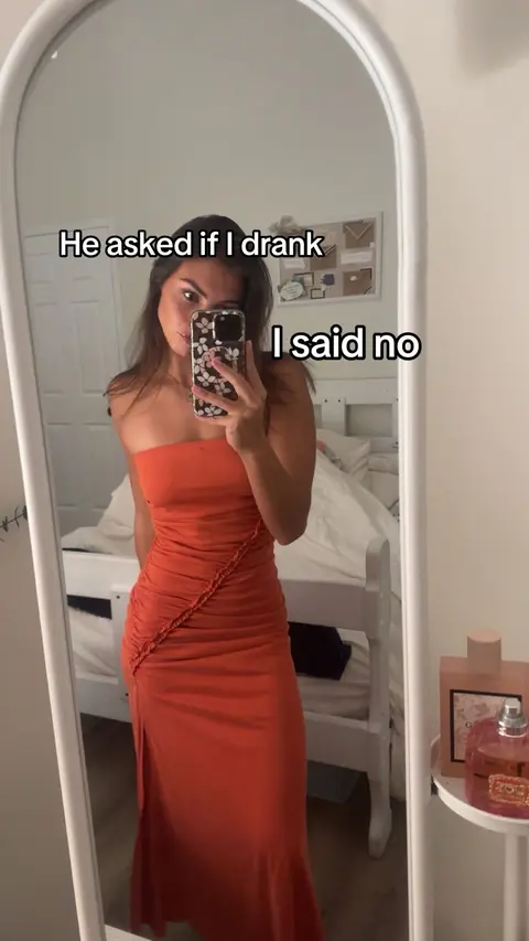 A woman is filmed from the chest up in a mirror, holding her phone up to take the selfie, with text overlaid on the top, the frame of the mirror is visible at the frame’s edge and the reflection of the room is visible in the mirror, which shows a bed and a table.
