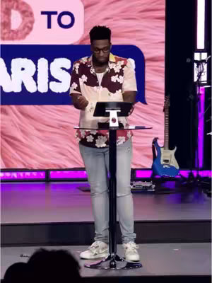 A man in a patterned shirt stands at a podium with a microphone, likely speaking at a church service. A blue guitar is in the background on the right side of the frame. The backdrop is brightly lit with pink and purple colors.