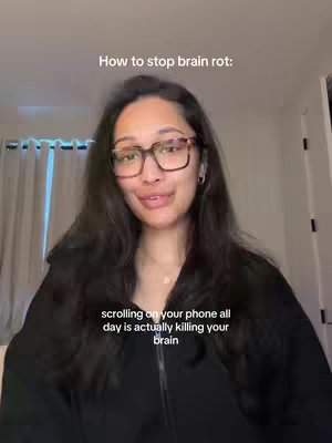 A woman with long, dark hair and glasses is centered in a video frame. Above her head, text reads 'How to stop brain rot:'. Below her, text says, 'scrolling on your phone all day is actually killing your brain'. She is wearing a dark top and has a neutral expression, partly visible in a room with a curtain.