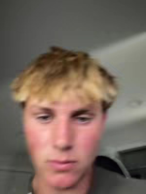 Close-up shot of a young man with light blond hair. The focus is on his face, which appears slightly blurred, giving the image a soft focus. The background is a blurred, neutral grey color.