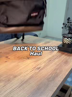 A wooden table is shown in the foreground, angled slightly downward. A dark-colored backpack, partially blocking the top of the frame. The lighting is bright, creating highlights on the wooden surface and the bottom of the backpack.Text overlayed on top of the image reads BACK TO SCHOOL Haul, and is set in a white font.