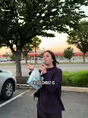 A woman stands in a parking lot with a microphone, speaking. Text overlays the image. Trees and a shopping center are in the background.