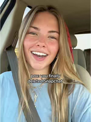 A young woman with blonde hair smiles widely while seated in a car. She is wearing a blue shirt and several necklaces. The background shows a car interior, with a seatbelt visible.
