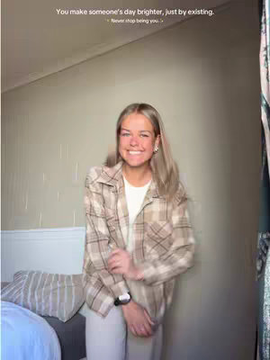 A young woman with blonde hair and a plaid shirt smiles broadly, posing in front of a neutral-toned wall. Light filters into the room, and a bed and some curtains are visible in the background.