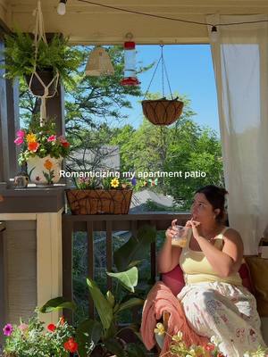 A woman sits in a cozy outdoor space, sipping from a glass. The scene is a balcony decorated to resemble a European summer setting. Overhead, string lights, hanging plants, and a bird feeder complete the ambiance. Colorful flowers are arranged in pots and baskets. The background reveals a clear blue sky and lush green trees, suggesting a sunny day.