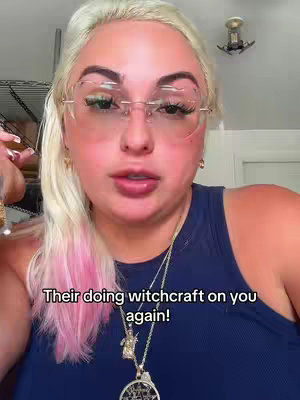 A woman with blonde hair and pink highlights in front, is wearing glasses and a dark blue top. The background is a room with a few visible design elements. Text overlayed on the cover says, 'Their doing witchcraft on you again!'