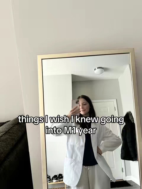 A person is taking a mirror selfie. The person is standing in front of a gold-framed mirror. The individual is wearing a white lab coat over a dark shirt and light pants. The person's reflection is visible. The background includes a door and part of a bed, with a few shoes visible below in the mirror.