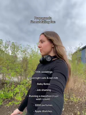 A young woman with long brown hair stands outdoors, looking to her left. She is wearing a black long-sleeved shirt and a black headphones around her neck. The background shows green foliage and a cloudy sky, with text overlays on the image.