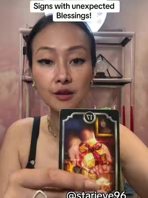 A woman with dark hair is holding a tarot card in front of her. The image includes a close-up of her face and the card with a blurred background, which could be a room.