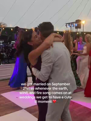 A couple is embracing on a dance floor inside a wedding reception. The bride is in a darker gown, and the groom is in a blazer. Pink and white square tiles make up the dance floor. There are other wedding guests in the background.