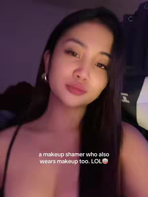 A selfie-style image of a woman with long, dark hair. She has fair skin and is wearing what appears to be a dark top, possibly a tank top or dress. The background is dimly lit in shades of purple. Text overlayed on the image says, "a makeup shamer who also wears makeup too. LOL" with a winking face emoji.