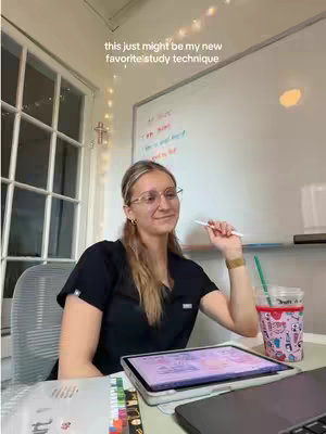 A young woman with blonde hair and glasses sits at a desk, smiling while holding a pen, with a laptop, a tablet, and a whiteboard behind her. A drink with a straw sits to her right, and a window with lights is visible. A text overlay at the top says, 'this just might be my new favorite study technique.'