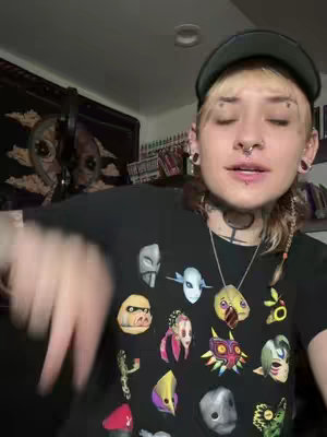 A young person with light skin and light-colored hair is centered in a medium shot. They wear a black graphic t-shirt with various colorful masks on it. They are wearing a grey cap, black tunnels in their earlobes, and a septum piercing. Their facial expression is neutral, with their eyes closed. They are looking down toward their lower left. The background shows a glimpse of a room with some decor.