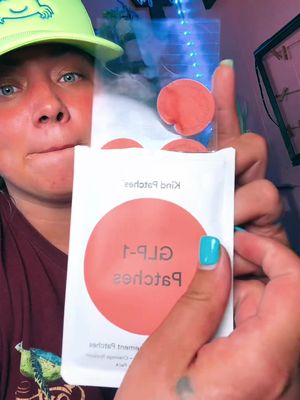 A woman in a maroon shirt holds a package of self-adhesive circular patches. The woman is looking at the camera, with an expression that appears to be a bit skeptical, and wearing a green cap. The package is white with a large orange circle and text indicating the patch type in the center. The woman's nails are painted bright blue. The background behind the woman has a faint colorful glow.